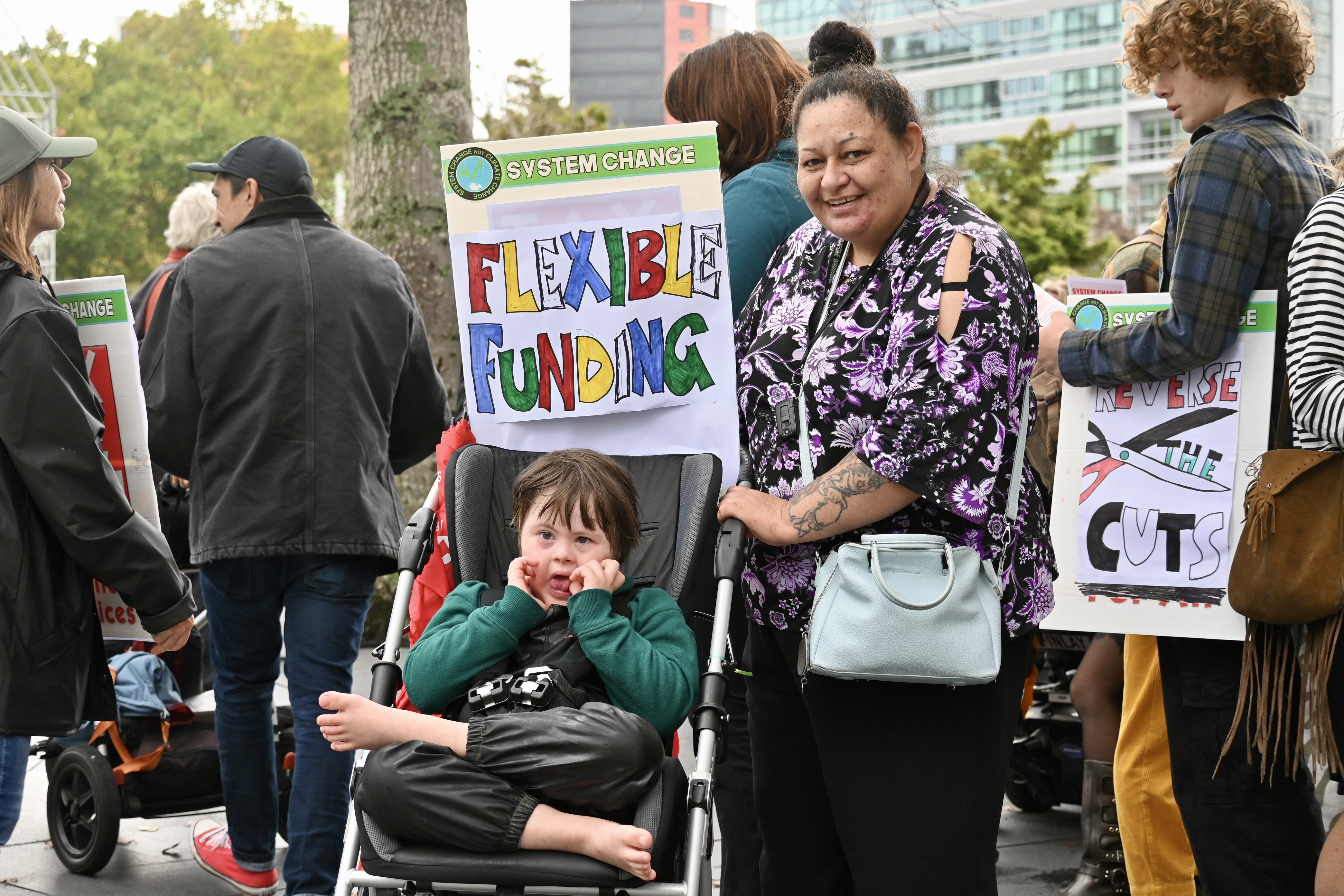 A child with Down syndrome sits in a pushchair and a woman stands beside him with a placard reading: System change. Flexible funding. 