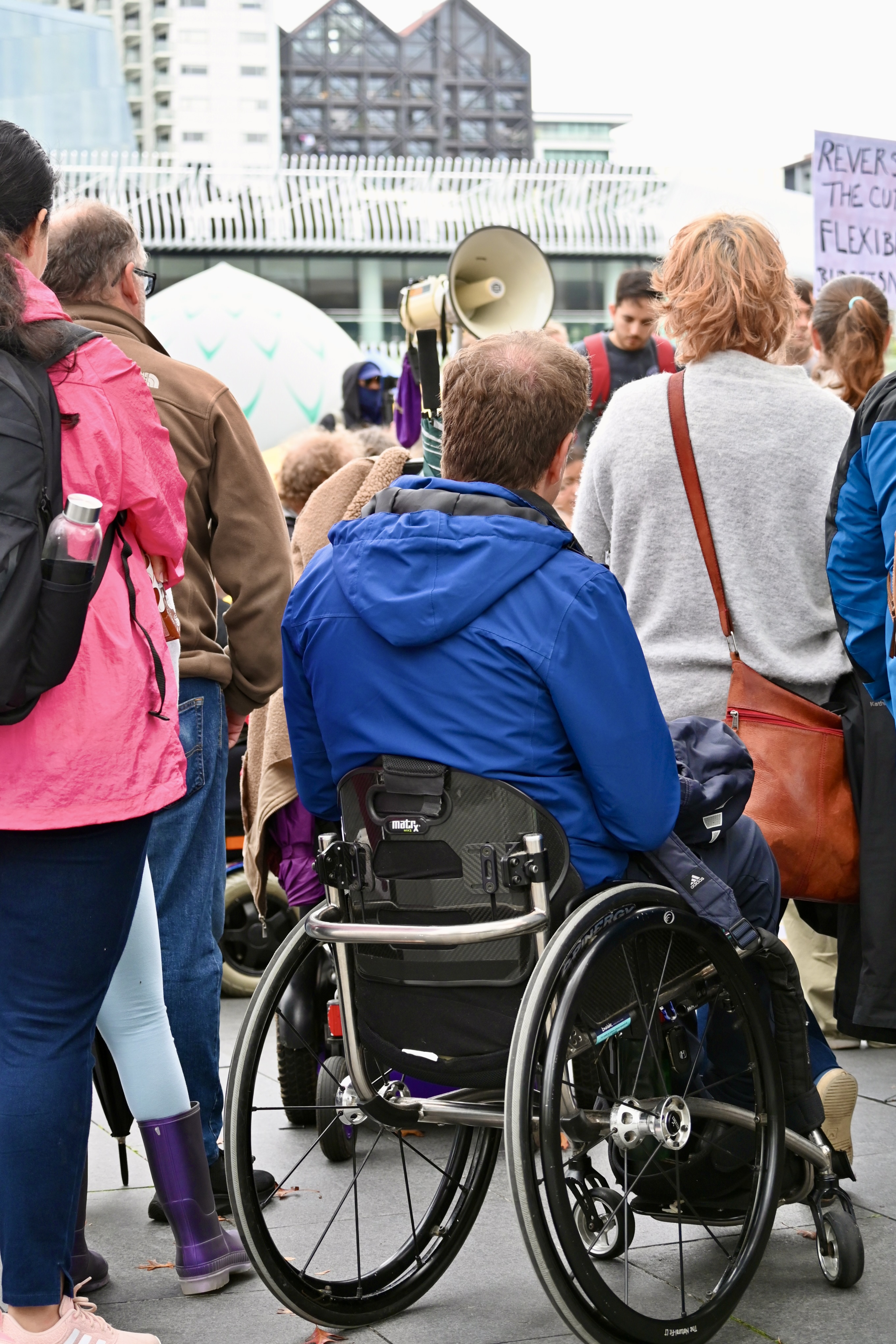 The back of a wheelchair-user wearing a blue jacket is among the crowd at a protest rally.