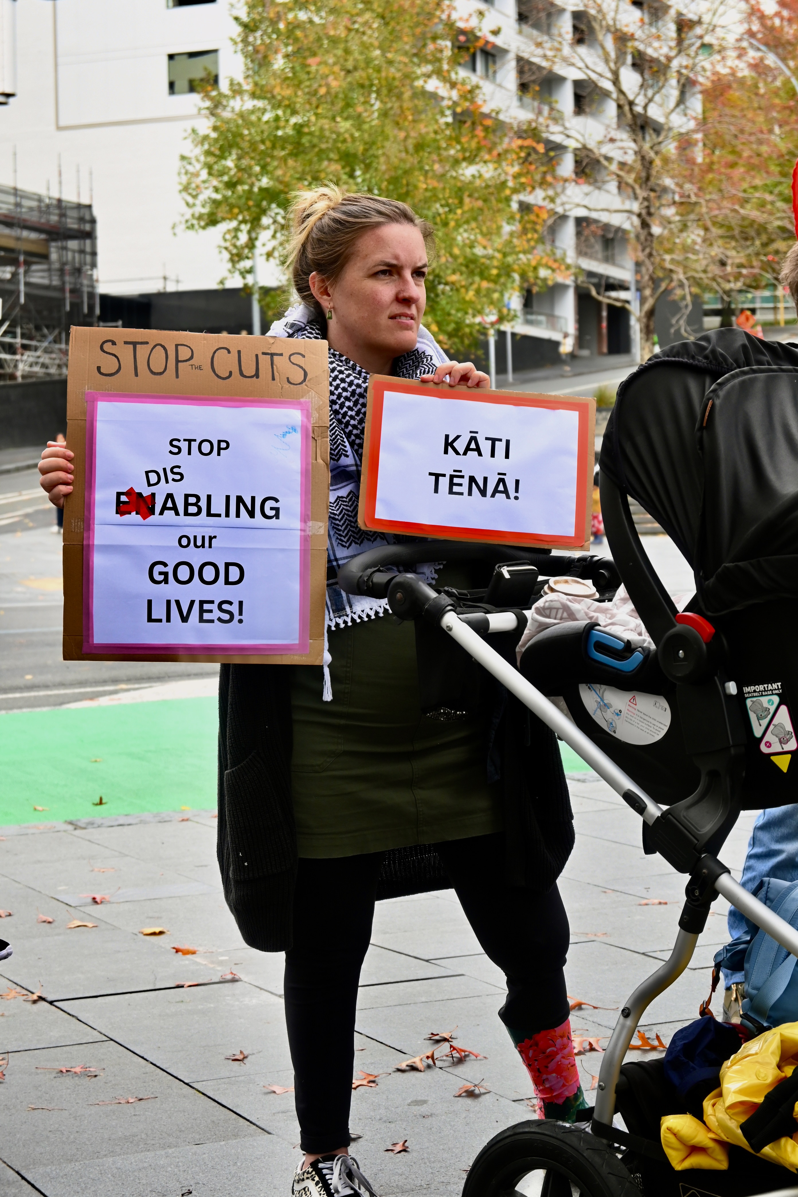 A woman with a prosthetic leg stands behind a baby pram and holds up two signs that read: Stop cuts, stop disabling our good lives! And: Kāti tēnā!