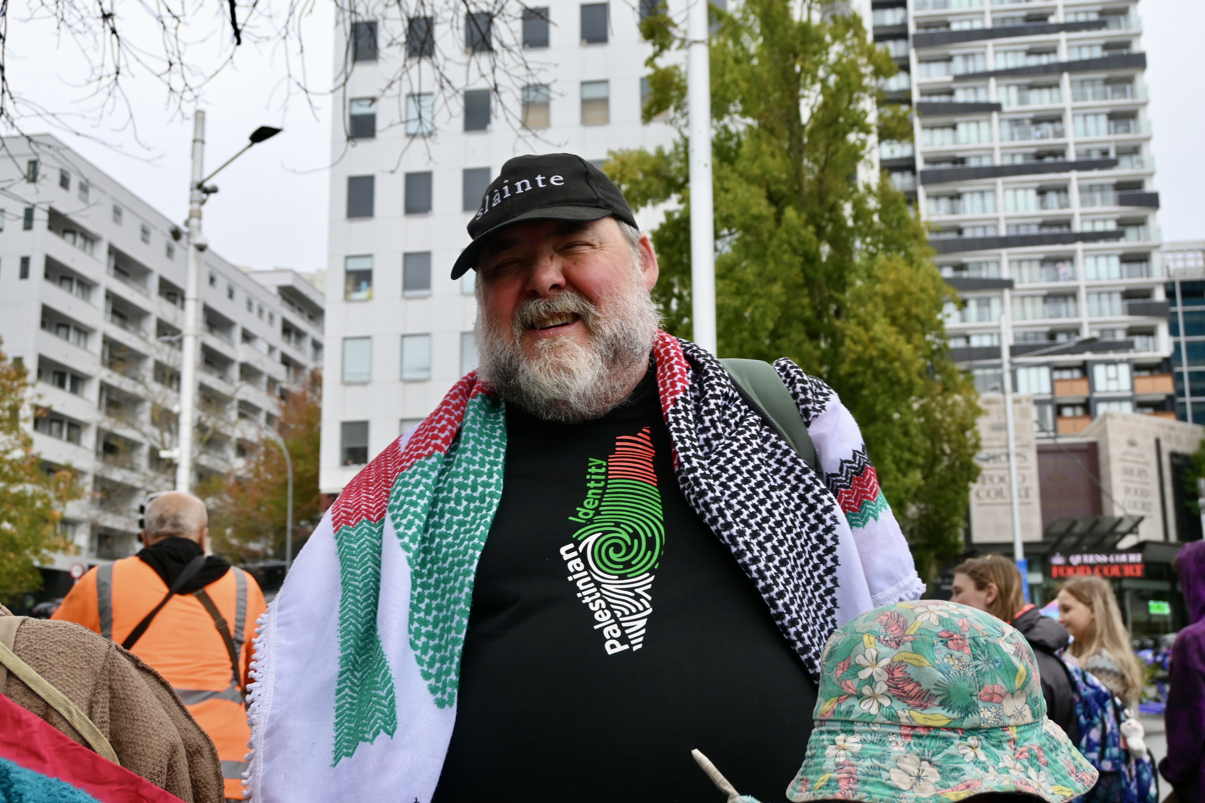 A man with a black cap and pro-Palestine clothing is at a protest in front of city buildings.