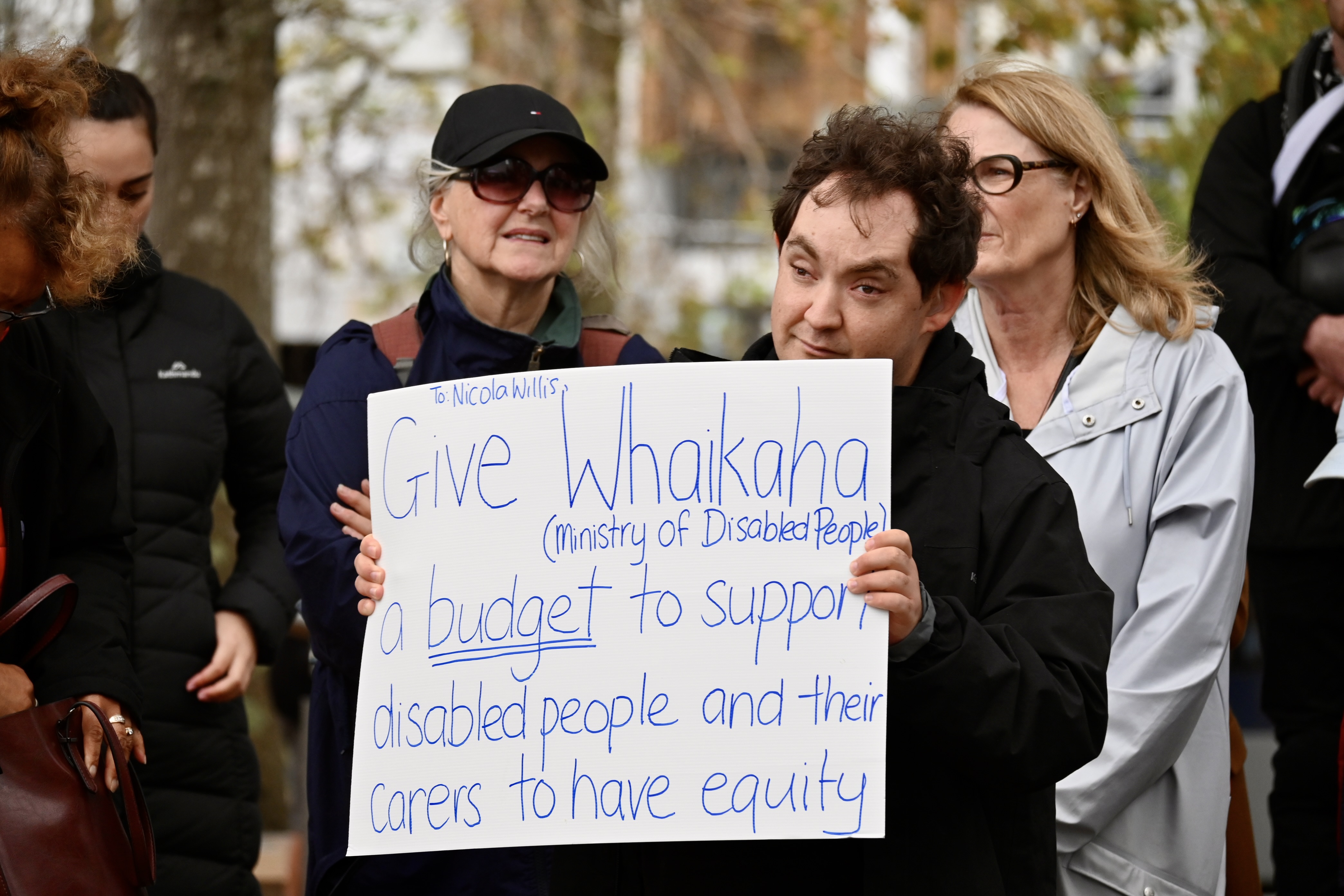 A man holds up a sign that reads: To Nicola Willis, Give Whaikaha (Ministry of Disabled People) a budget to support disabled people and their carers to have equity.